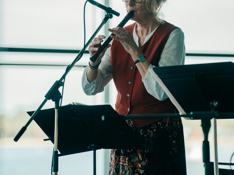 Image of An older woman wearing glasses and a red waistcoat plays a recorder into a microphone, standing by a music stand with sheet music in front of large windows overlooking water.