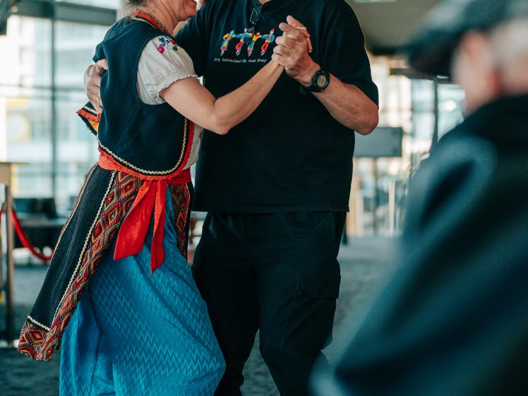 Image of A smiling woman in traditional clothing and a man in black attire dance together indoors, holding hands and looking at each other. Another person is partially visible in the foreground.