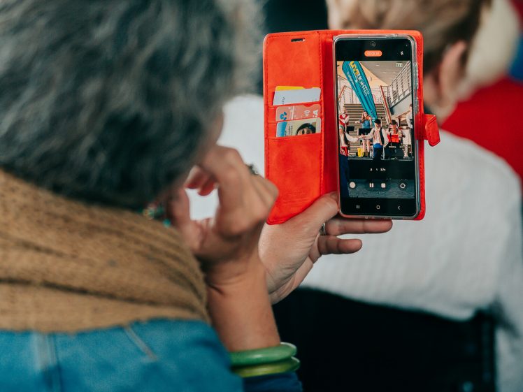 Image of A person with grey hair and a brown scarf holds up a mobile phone in a red case, taking a photo or recording an event. The phone screen shows a group of people and colourful banners. The person is seated among others.