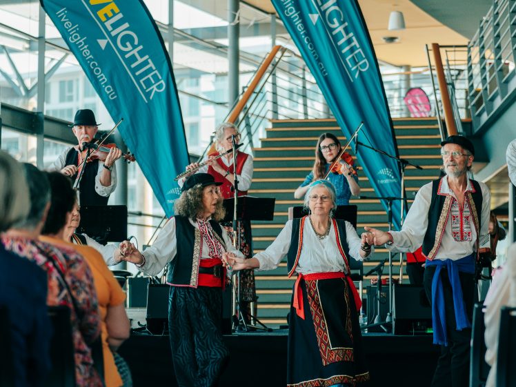 Image of A group of people in traditional costumes dance whilst musicians play onstage behind them at an indoor event with LiveLighter banners and seated audience members watching.