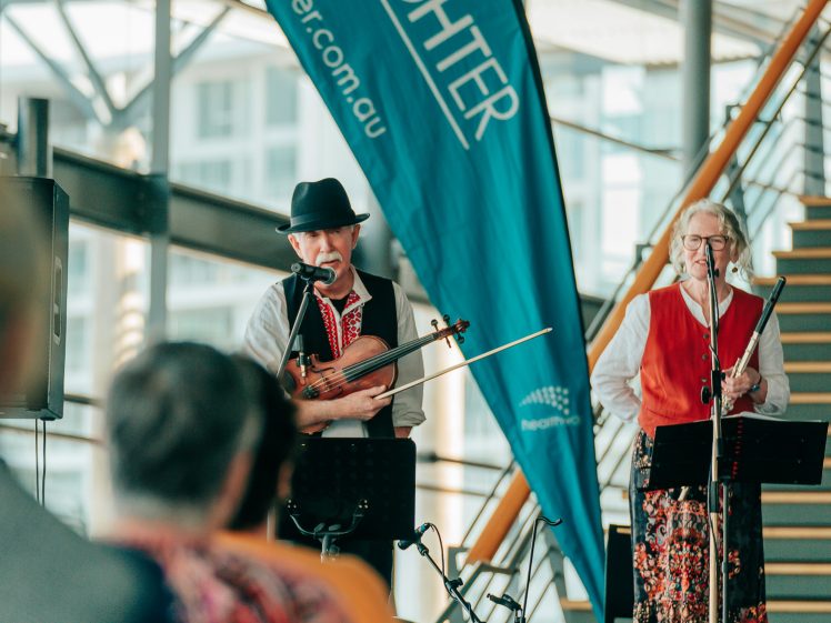 Image of Two musicians perform indoors beside a blue LiveLighter banner. One plays the violin and sings into a microphone; both wear folk-style clothing. Audience members are visible in the foreground.