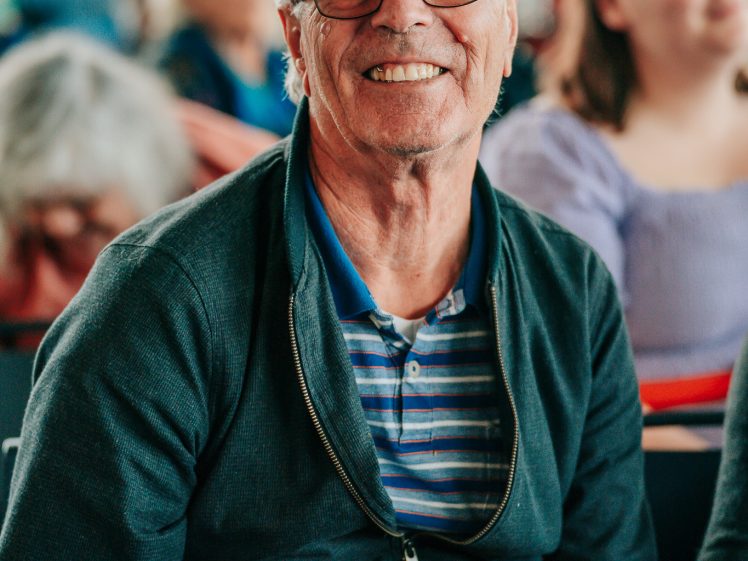Image of An older man with grey hair and glasses, wearing a striped shirt and dark jacket, sits smiling in a crowded indoor setting with other people in the background.