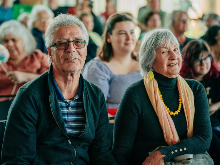 Image of An older man and woman sit smiling in the foreground at an indoor event, with other people seated and blurred in the background. The man wears glasses and a dark jacket; the woman wears a scarf and a yellow necklace.