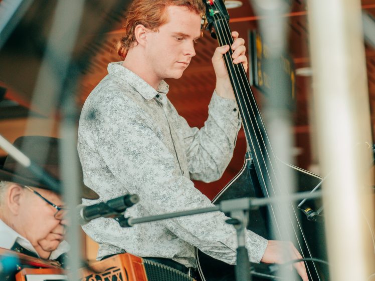 Image of A young man with reddish hair plays a double bass on stage, while an older man beside him plays an accordion. Microphones and musical equipment are visible in the foreground.