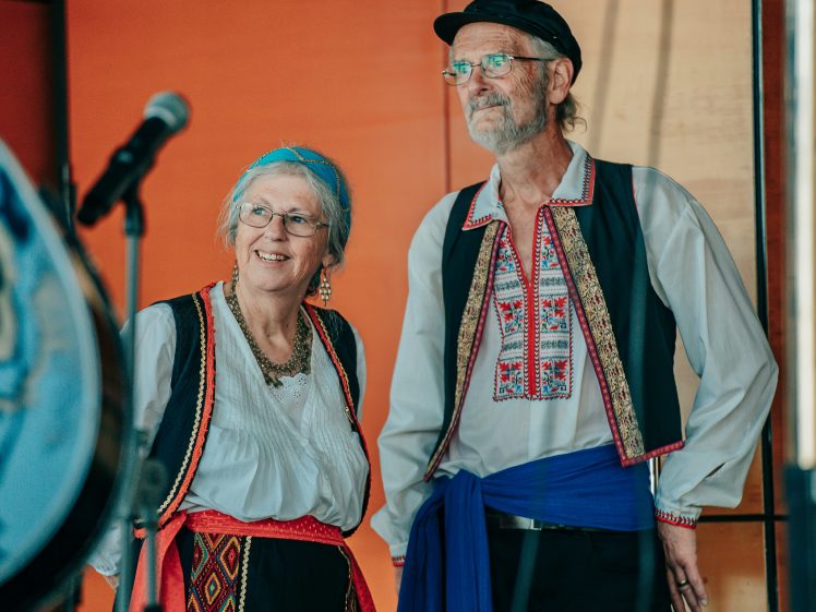 Image of Two older adults in traditional folk costumes stand side by side, smiling. Both wear embroidered shirts, waistcoats, and sashes; the woman has a headscarf and jewellery, while the man wears a cap. A microphone is visible in the foreground.