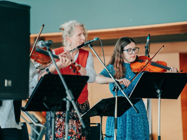 Image of Two women play violins on stage behind music stands and microphones. The older woman has white hair and wears a red waistcoat; the younger woman has long brown hair, glasses, and a blue dress. A loudspeaker is visible in the foreground.