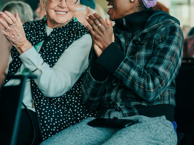 Image of Two women, one older and one younger, sit side by side smiling and applauding. The older woman wears glasses and a spotted waistcoat, while the younger woman wears a purple headscarf and checked jacket. Both appear to be enjoying an event.