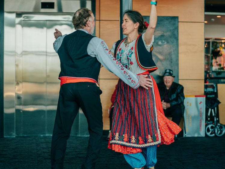 Image of A man and a woman in traditional folk costumes dance together indoors, smiling and holding hands, whilst people sit and watch in the background.