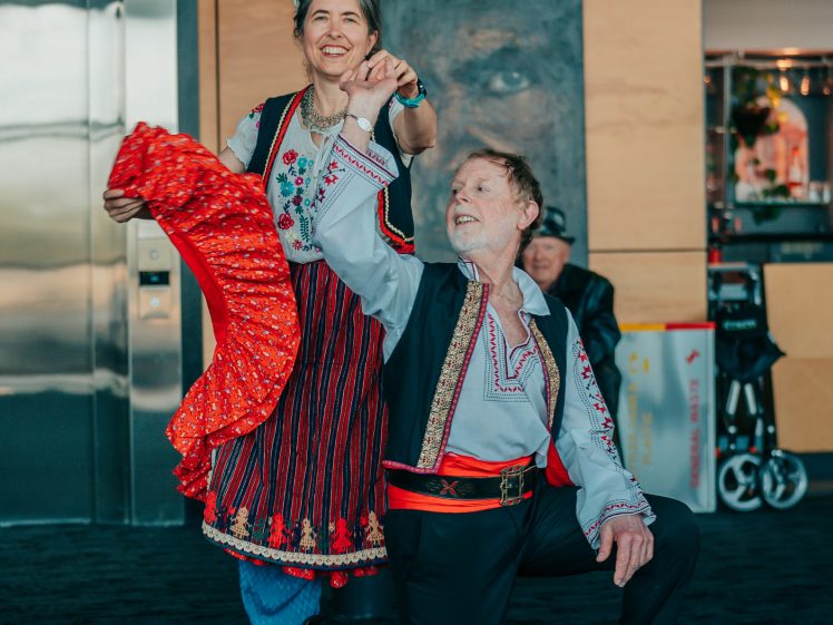 Image of An older man and woman perform a traditional dance indoors, wearing colourful folk costumes. The man kneels on one knee, holding the woman's hand, while she holds a bright red scarf and smiles.