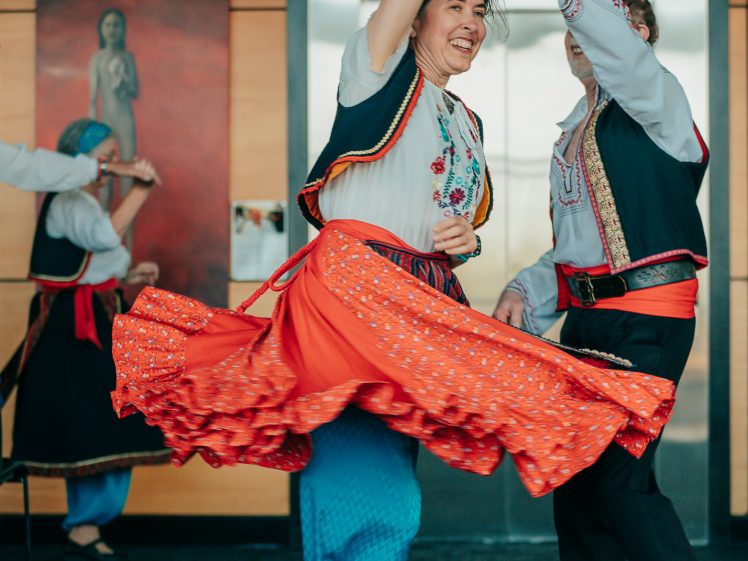 Image of A woman in colourful traditional folk attire smiles as she twirls, holding hands with a male partner in matching costume. Another pair, also in folk dress, appears in the background, blurred.