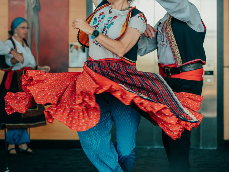 Image of Two people in colourful traditional clothing joyfully dance indoors. The woman’s red skirt twirls as she spins, and both wear embroidered shirts. Other dancers and a painting are visible in the background.