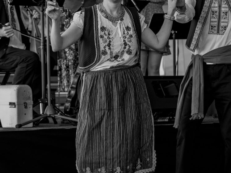 Image of A woman in traditional folk clothing dances and smiles, surrounded by musicians and other performers in similar attire, during a lively cultural event. The image is in black and white.