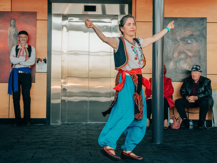 Image of An older woman in colourful traditional clothing dances with arms raised in front of a lift, while three seated people and one standing man watch. Two large portrait paintings hang on the wall behind them.