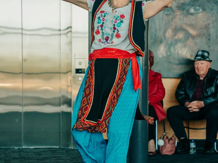 Image of A woman in colourful traditional clothing dances indoors, smiling with arms raised. She wears a floral top, blue trousers, red sash, and brown shoes. Seated people and a painting appear in the background.