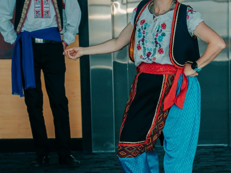 Image of A woman and a man wear traditional folk costumes. The woman smiles and gestures while dancing, wearing blue trousers, a white embroidered blouse, and a black waistcoat. The man stands in the background, watching her, dressed in similar attire.