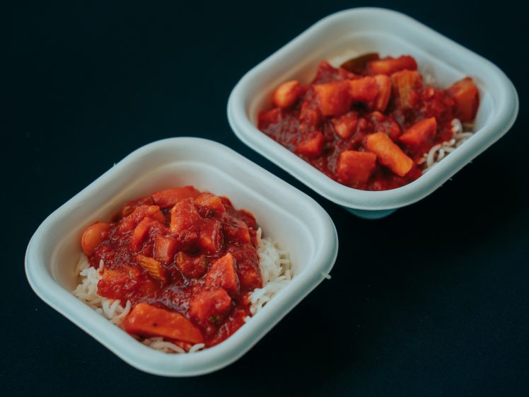 Image of Two square white bowls filled with white rice topped with a chunky red tomato-based stew containing vegetables, placed on a dark surface.