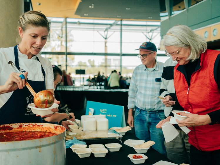 Image of A woman in a white shirt and apron serves soup from a large pot to an older woman in a red waistcoat, while two men wait nearby at a food table in a bright, indoor setting.