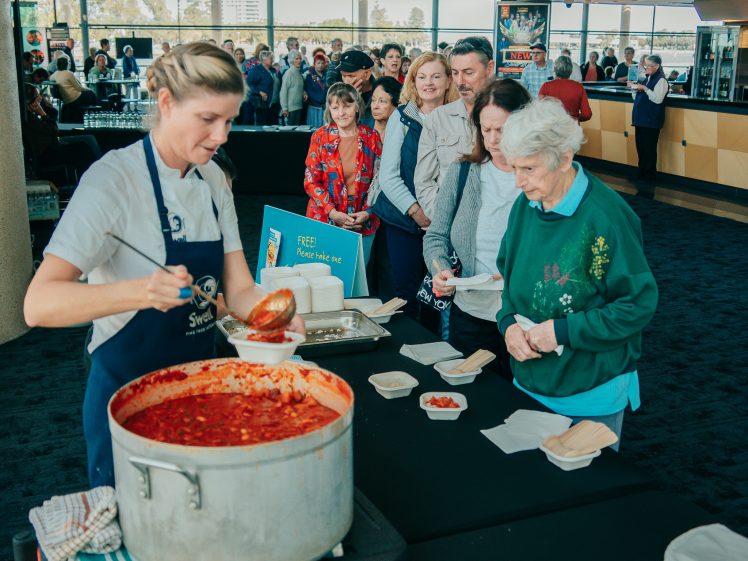 Image of A woman serves food from a large pot of red stew into bowls while a queue of people, some elderly, wait to be served at an indoor event. The atmosphere appears casual and friendly.