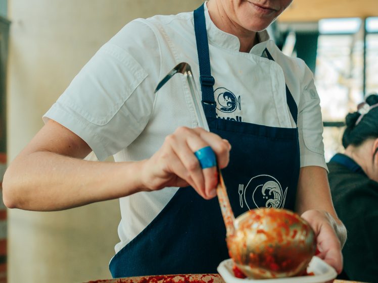 Image of A woman in a white shirt and blue apron ladles vegetable soup from a large saucepan into a bowl, focussed on her task.