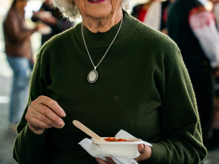 Image of An elderly woman with white hair and glasses, wearing a green jumper and a pendant necklace, smiles whilst holding a bowl of food and a wooden spoon. Blurry people are visible in the background.