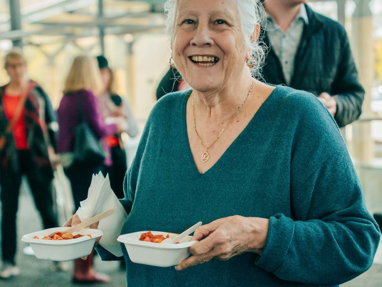 Image of An older woman with grey hair, wearing a blue jumper, smiles while holding two bowls of food and serviettes. Several people are seen in the blurred background indoors.