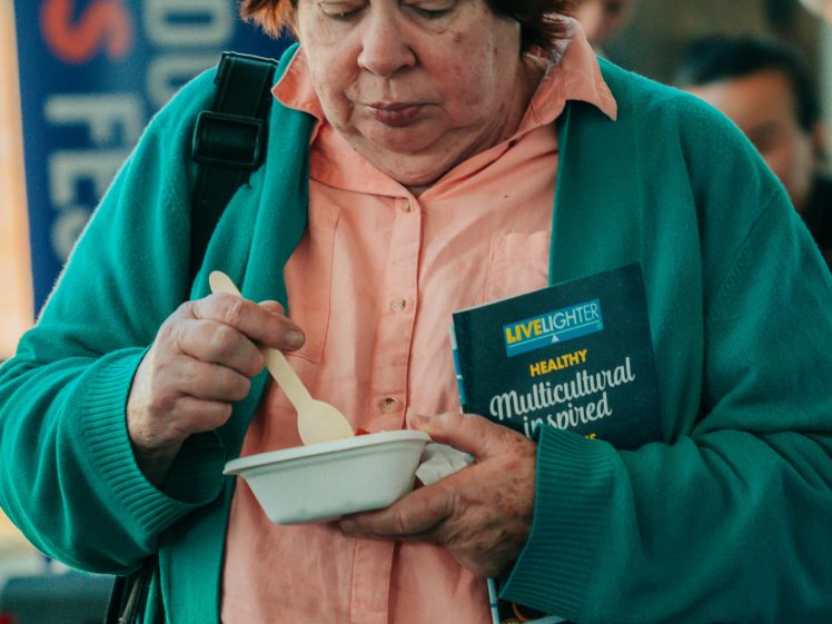 Image of An older woman in a green cardigan holds a bowl and eats with a spoon. She also holds a LiveLighter Multicultural flyer and stands indoors, with a colourful festival banner visible in the background.