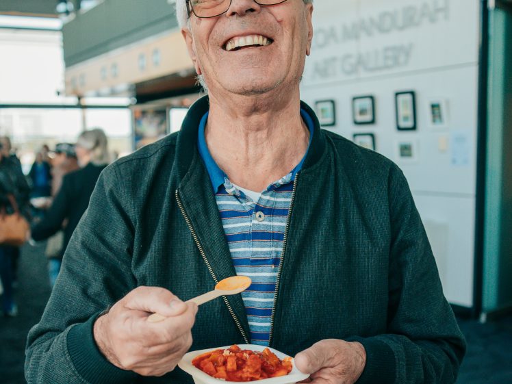 Image of An older man with grey hair and glasses smiles whilst holding a bowl of food and a spoon, standing indoors in a gallery space with framed artwork on the wall behind him.