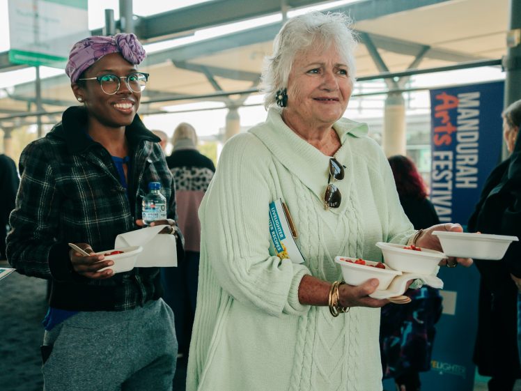 Image of Two women stand together holding food containers and spoons, smiling. The woman on the right has white hair and wears a light-green jumper; the woman on the left wears glasses, a purple headscarf, and a checked jacket. A festival sign is in the background.