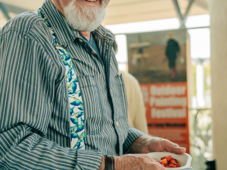 Image of An older man with a white beard, wearing a striped shirt, colourful braces, and jeans, holds a small tray of food and smiles at the camera indoors.