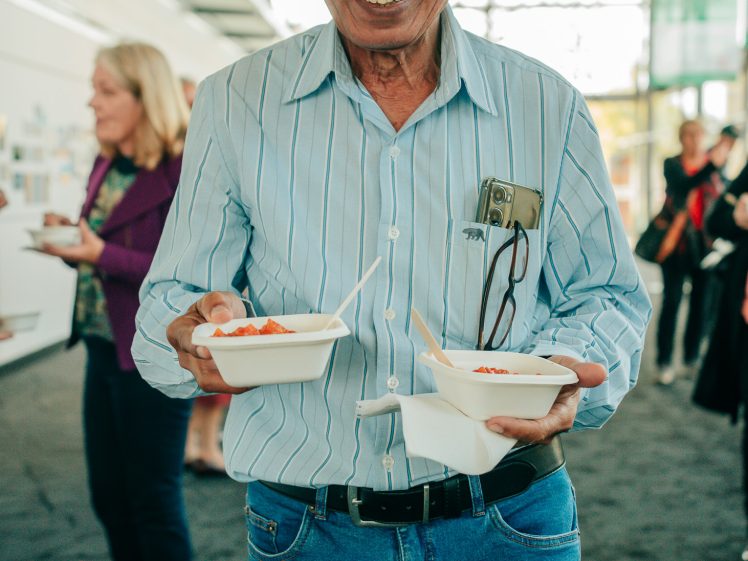 Image of An older man in a striped shirt and blue jeans smiles while holding two bowls of food. A mobile and spectacles are in his shirt pocket. People and a bright, modern indoor space are visible in the background.