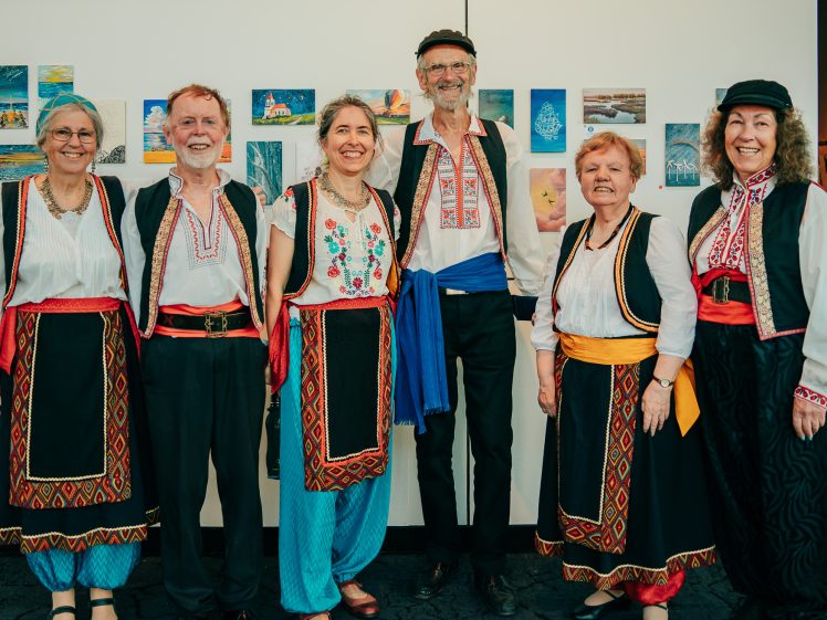 Image of Six smiling adults in traditional folk costumes stand side by side in front of a wall with small colourful artworks. The group includes three men and three women wearing embroidered shirts, waistcoats, and sashes.