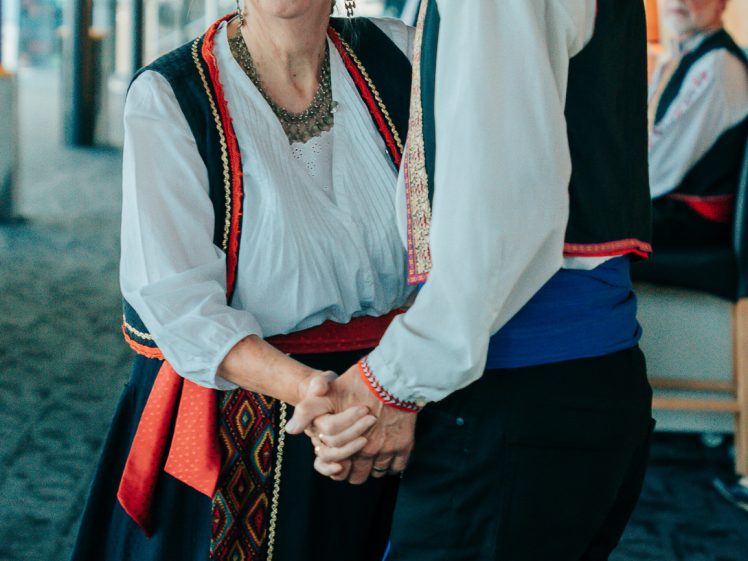 Image of An older couple in traditional folk costumes dance together indoors, smiling at each other. The man wears a waistcoat, hat, and sash; the woman wears a headscarf, necklace, and embroidered dress.