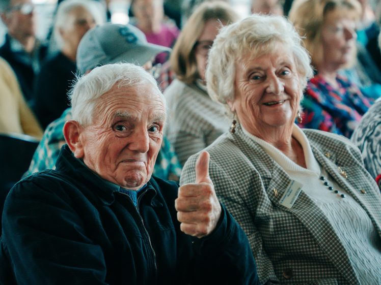 Image of An older man gives a thumbs up and smiles, sitting next to an older woman who is also smiling. Both are in a crowd with several people in the background.
