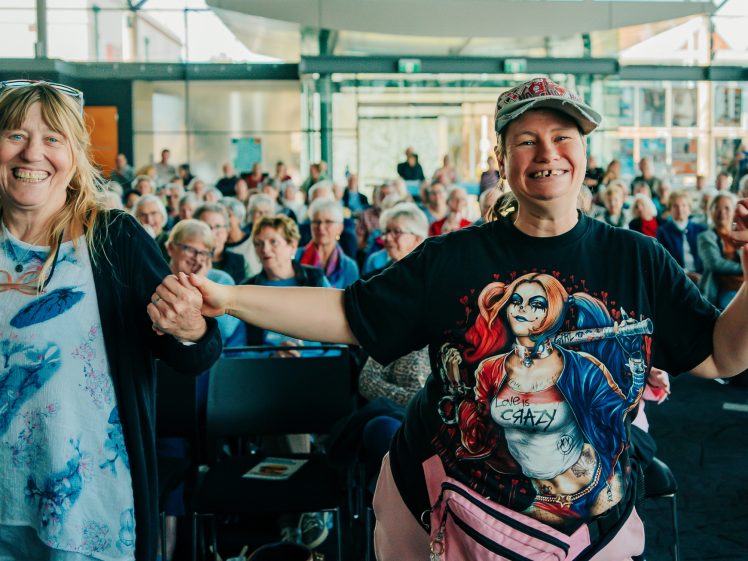 Image of Two smiling women stand at the front of a crowded room, holding hands. One wears a Harley Quinn T-shirt and a cap, while the other wears glasses and a cat-themed top. Many people sit in rows behind them. The mood is cheerful.