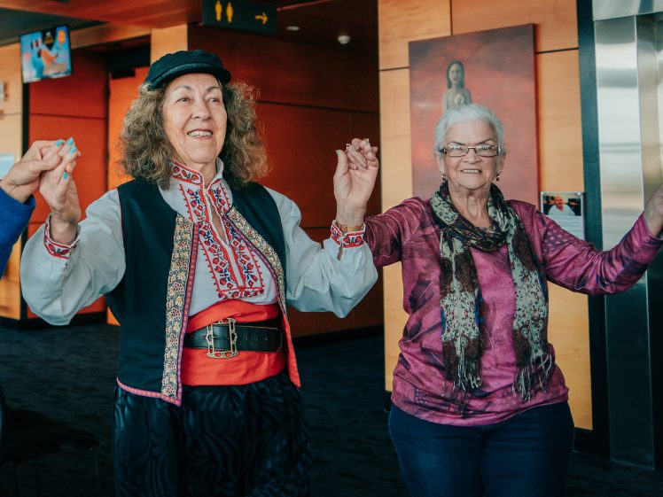 Image of Two older women smiling and holding hands, standing indoors. One wears traditional folk clothing with embroidery and a black cap, while the other wears glasses, a pink top, and a scarf. They appear to be participating in a group activity.