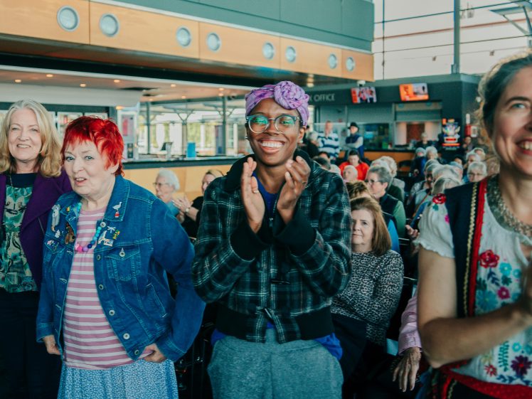 Image of A diverse group of people stands and smiles, some applauding, in a bright indoor space with many seated in the background, suggesting they are enjoying an event or performance.