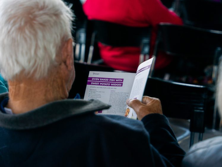 Image of An older person with white hair reads a recipe booklet, open to a page titled Oven-Baked Fish with Sweet Potato Wedges, while sitting among rows of black chairs at an indoor event.