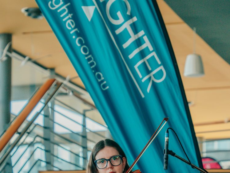 Image of A woman in a blue dress plays the violin indoors near a LIVELIGHTER promotional banner and a staircase. She wears glasses and stands in front of a microphone.