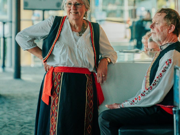Image of An older woman and man wear traditional folk costumes. The woman stands smiling with one hand on her hip, whilst the man sits nearby. The setting appears to be indoors, possibly at a cultural gathering or event.