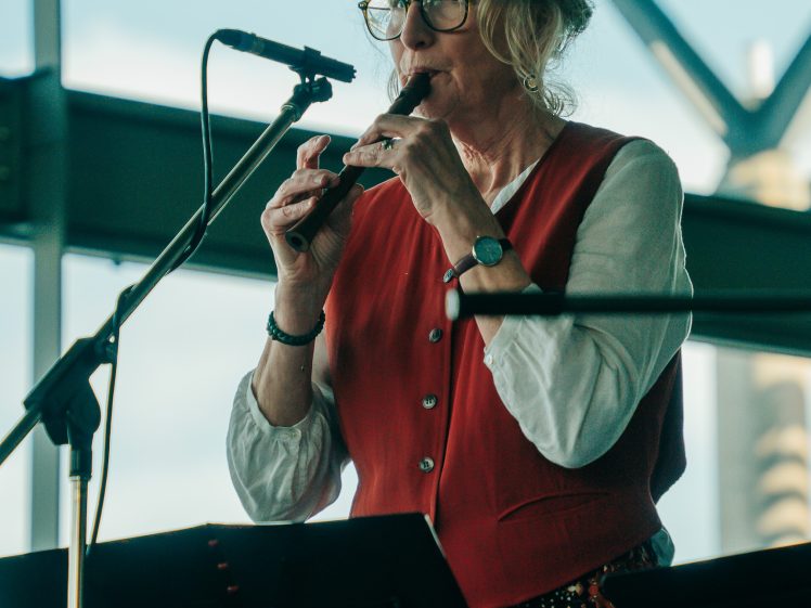 Image of An older woman with glasses and a red waistcoat plays a woodwind instrument in front of a microphone, with music stands and windows in the background.