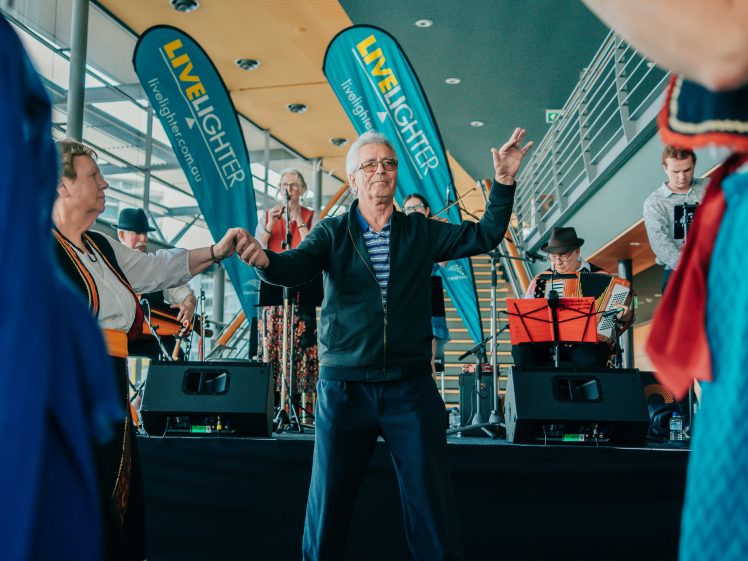 Image of An older man dances with others in front of a live band on stage. LiveLighter banners are in the background. People are enjoying a lively indoor event.