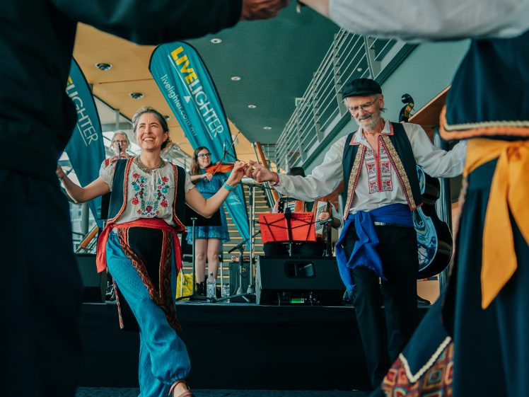 Image of A group of people in traditional folk costumes dance in a circle at an indoor event, smiling and holding hands, with musicians performing on a stage in the background.