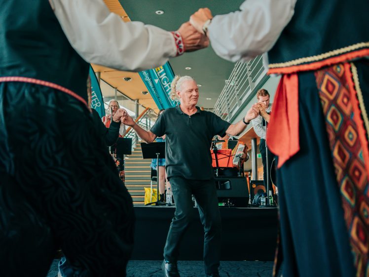 Image of An older man in a black shirt dances in a circle, holding hands with others in colourful traditional clothing. A band plays music in the background at an indoor event.