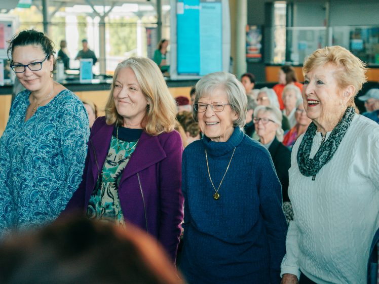 Image of Four smiling women stand together in a busy indoor setting, with other people seated in the background. The women appear to be enjoying themselves, dressed in casual, colourful clothing.
