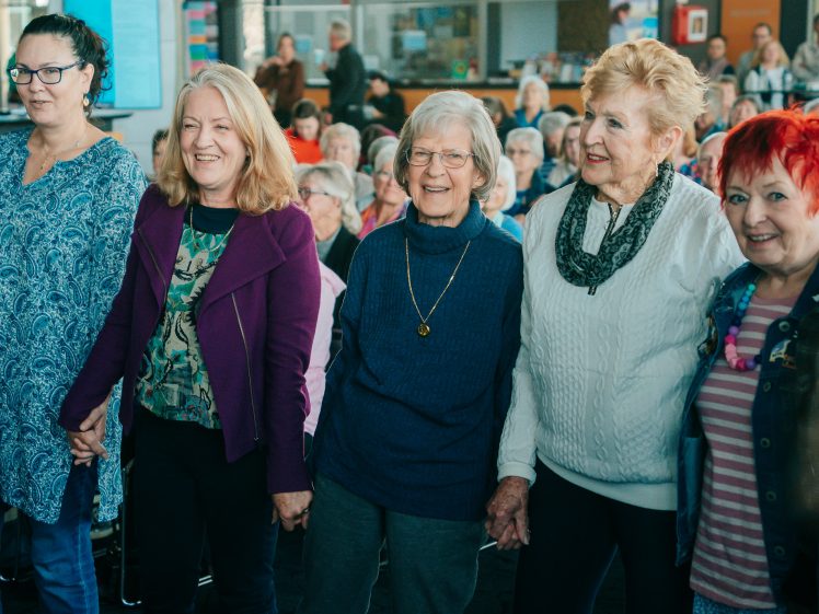 Image of A group of five older women stand together holding hands and smiling at an indoor event with an audience seated behind them.