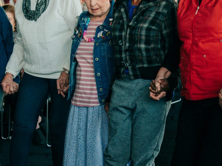 Image of A group of four women of different ages, holding hands and smiling as they dance together indoors. They are dressed in colourful, casual clothing and appear to be enjoying the moment.
