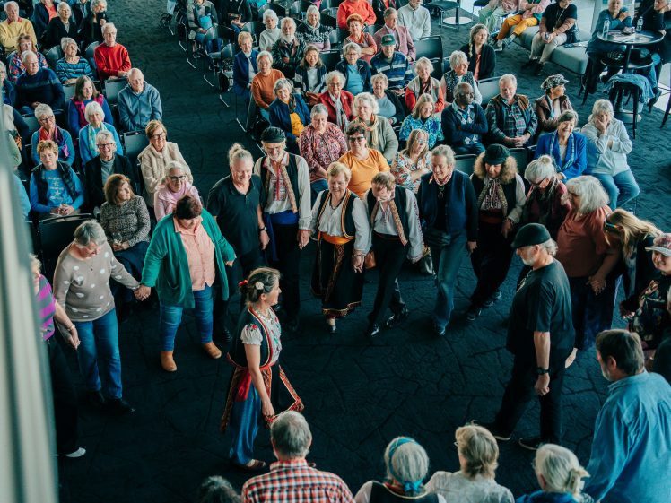 Image of A large group of people, some in traditional clothing, hold hands and form a circle while dancing indoors. Many seated spectators watch the dance, creating a lively and festive atmosphere.