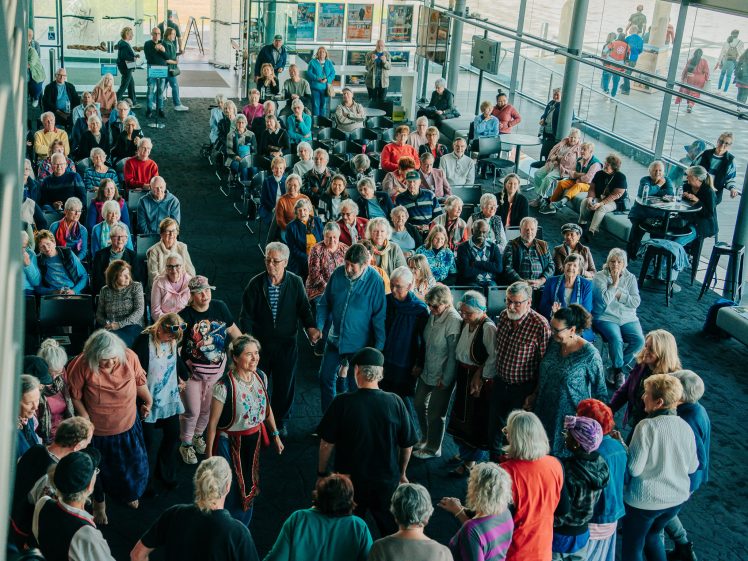 Image of A large group of older adults gathers in a spacious, bright indoor area, some seated and others standing in a circle, engaging in a communal activity or event. The atmosphere appears lively and sociable.