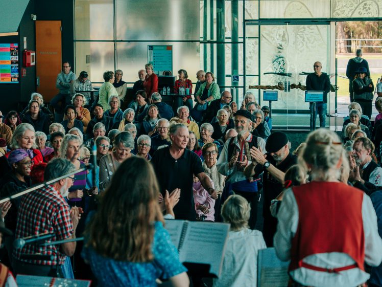 Image of A large group of people, mostly pensioners, gather indoors in front of floor-to-ceiling windows. Musicians perform to the left while the audience applaud and watch attentively in a brightly lit, spacious venue.