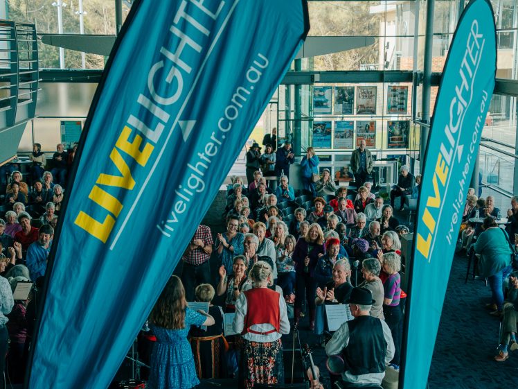 Image of A large crowd sits indoors facing a stage, framed by two tall blue LiveLighter banners. Musicians perform in front of the audience, and sunlight streams through large windows in the background.